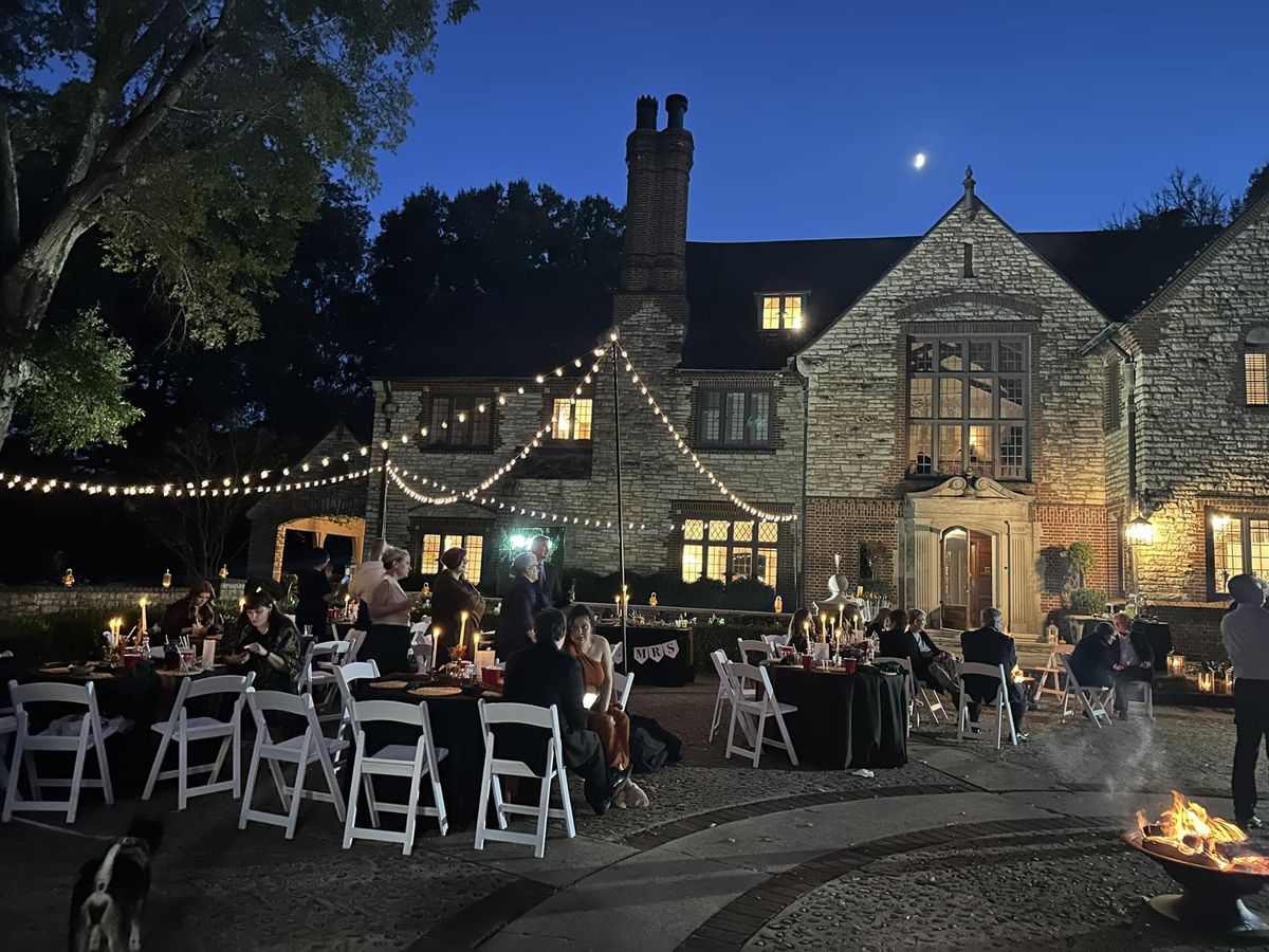 Evening outdoor wedding reception in front of stone mansion with string lights, candlelit tables, fire pit