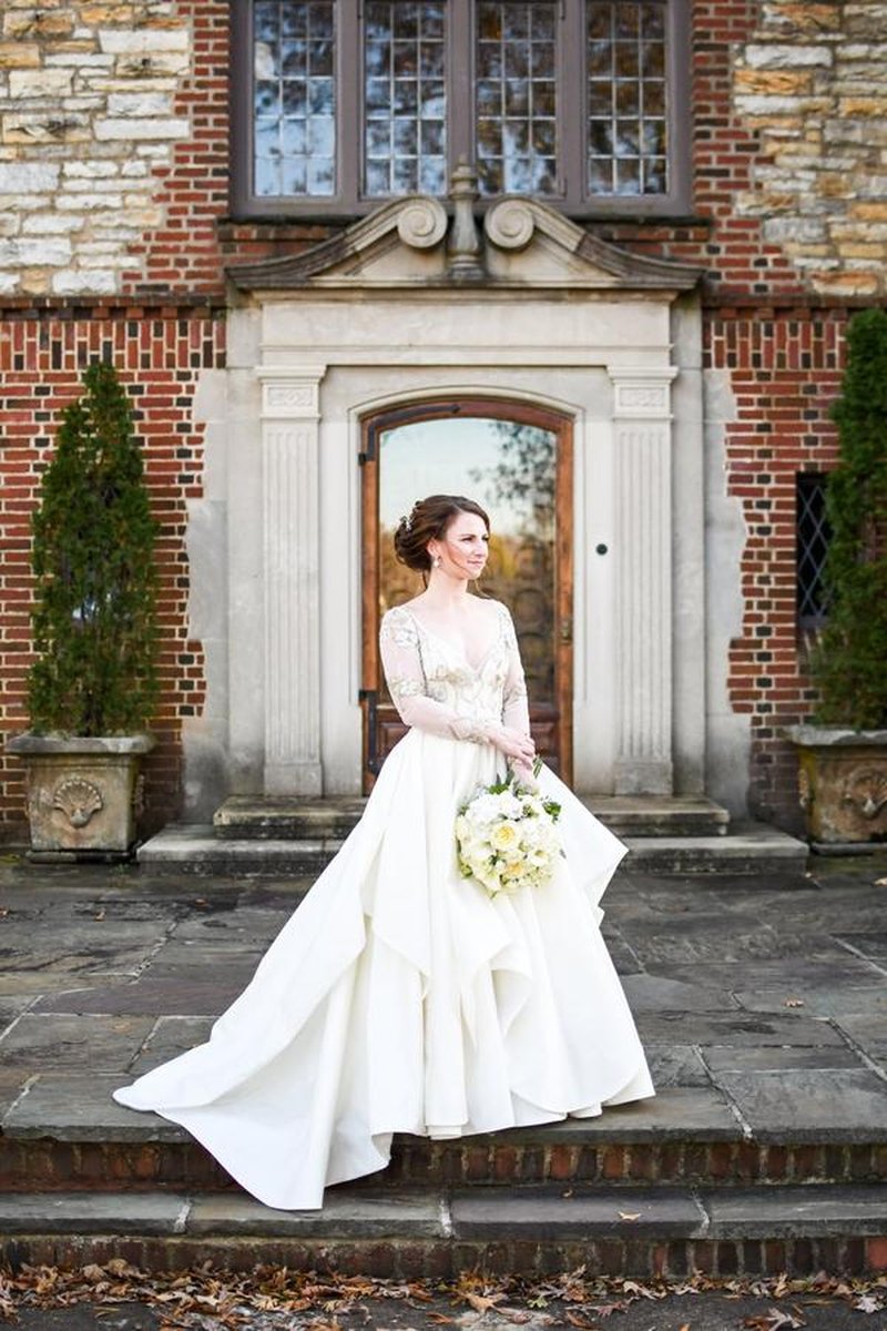 Bride in elegant long-sleeve beaded gown holding white bouquet before ornate mansion entrance