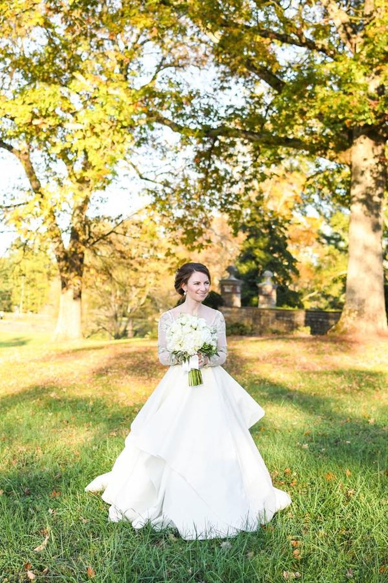 Bride holding white bouquet on estate lawn with autumn foliage and stone gate