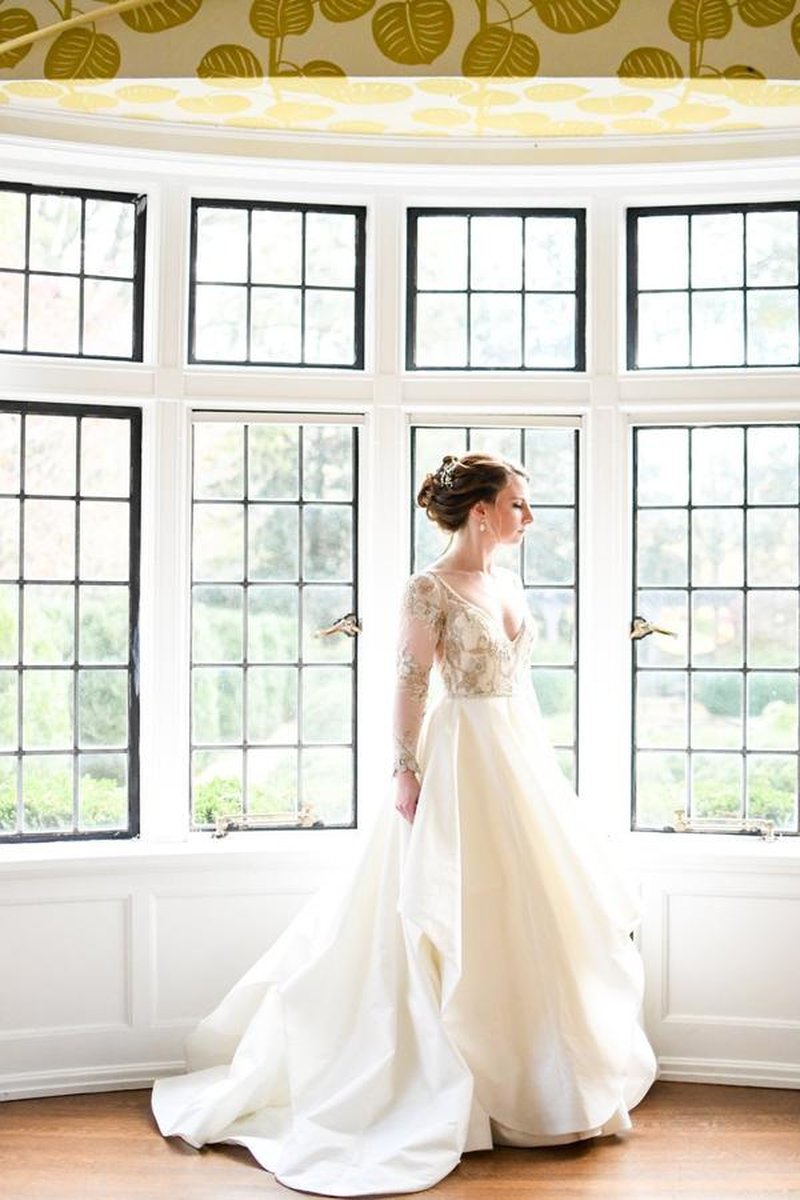 Bride standing in bay window alcove with leaded glass panes and botanical wallpaper ceiling