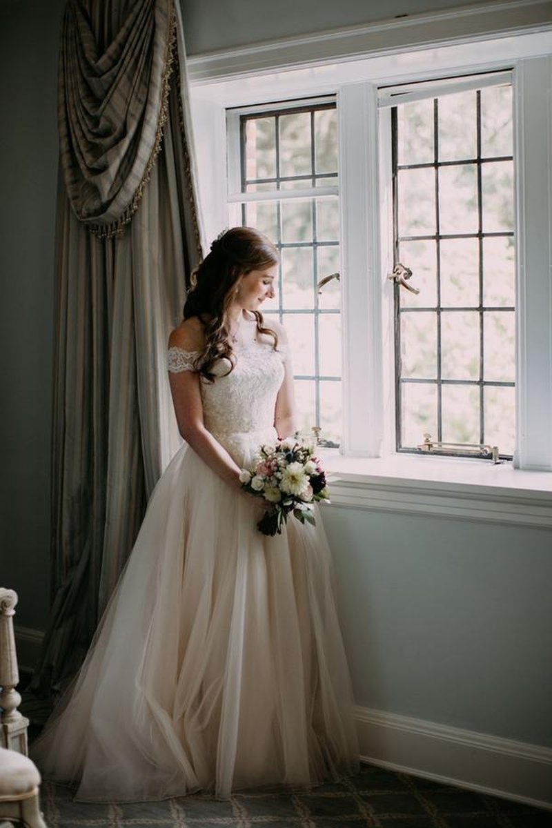 Bride holding bouquet by leaded glass window with elegant drapes in mansion room