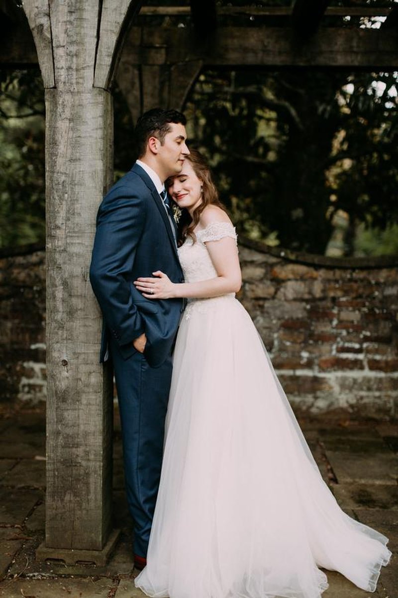 Bride and groom embracing under rustic wooden pergola with aged brick wall backdrop