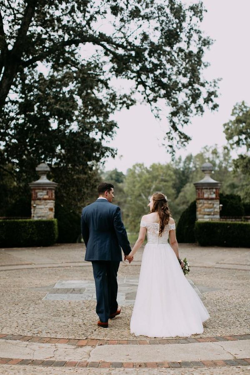 Bride and groom walking hand-in-hand away from camera through estate stone pillars