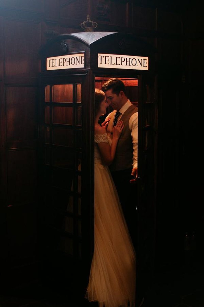 Bride and groom embracing inside vintage British telephone booth in wood-paneled room