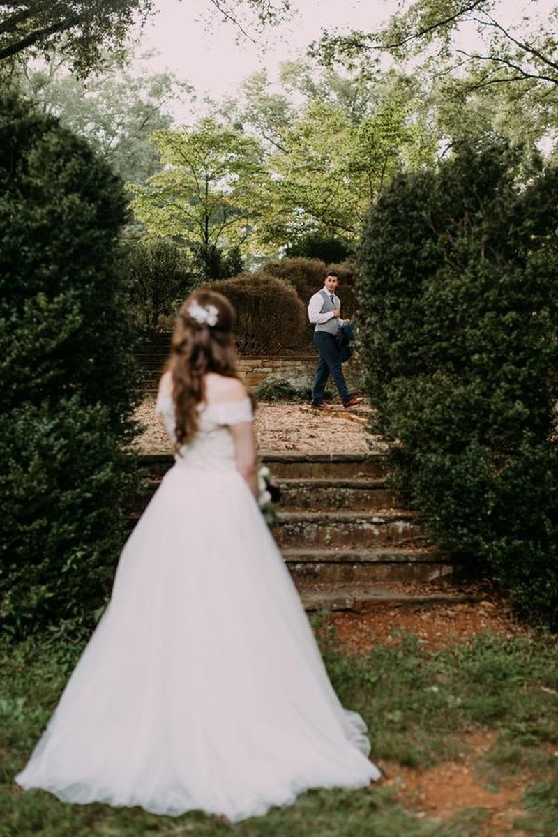 Bride in white gown watches groom approach through lush garden pathway with stone steps