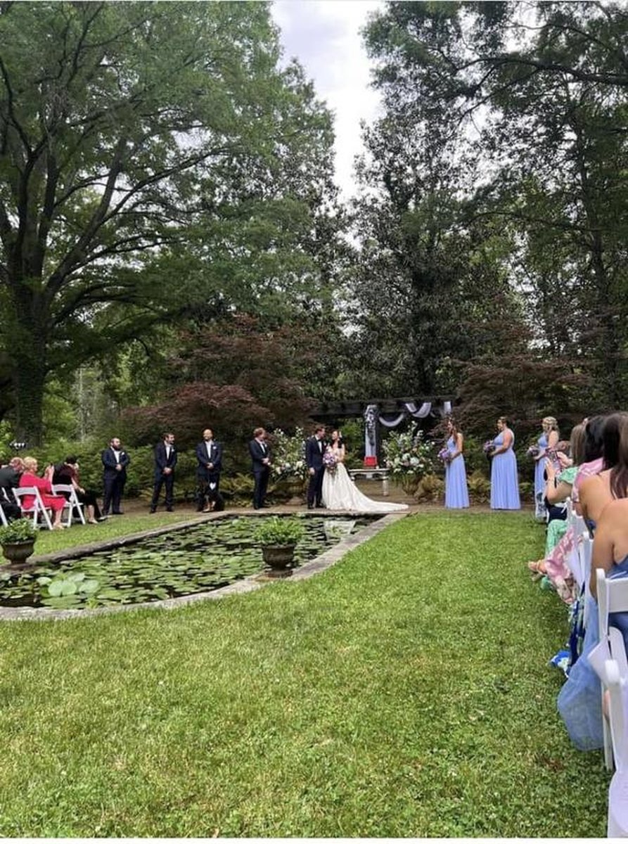 Outdoor wedding ceremony by lily pond with bridal party and guests seated under tall trees