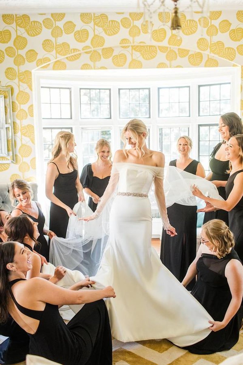 Bridesmaids in black dresses gathered around bride helping with veil and dress in elegant room