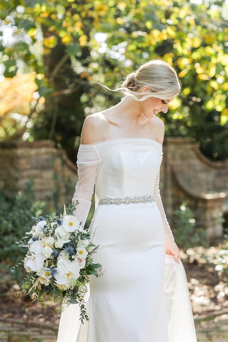 Bride smiling in off-shoulder gown holding white and blue bouquet in garden