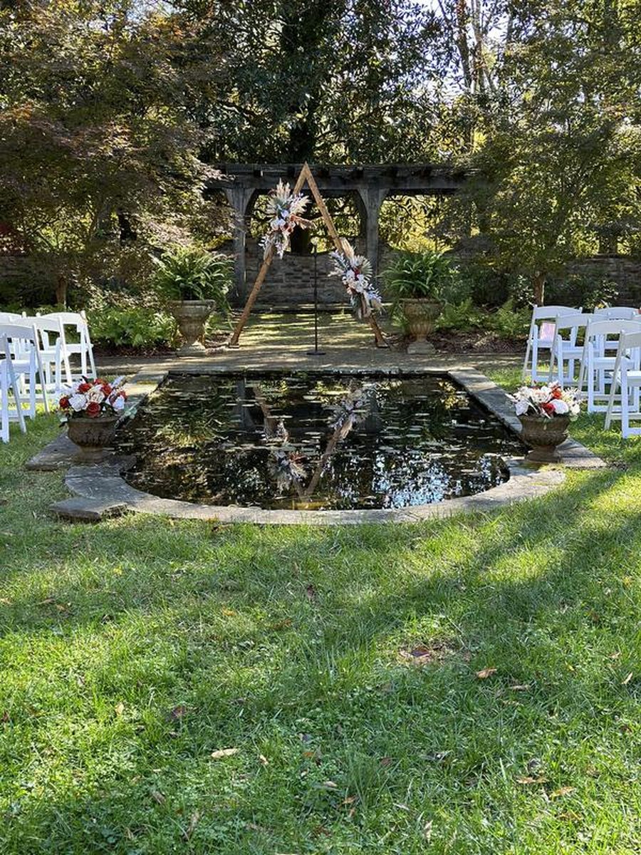 Outdoor wedding ceremony setup with triangular wooden arch, reflecting pond, floral arrangements, white chairs