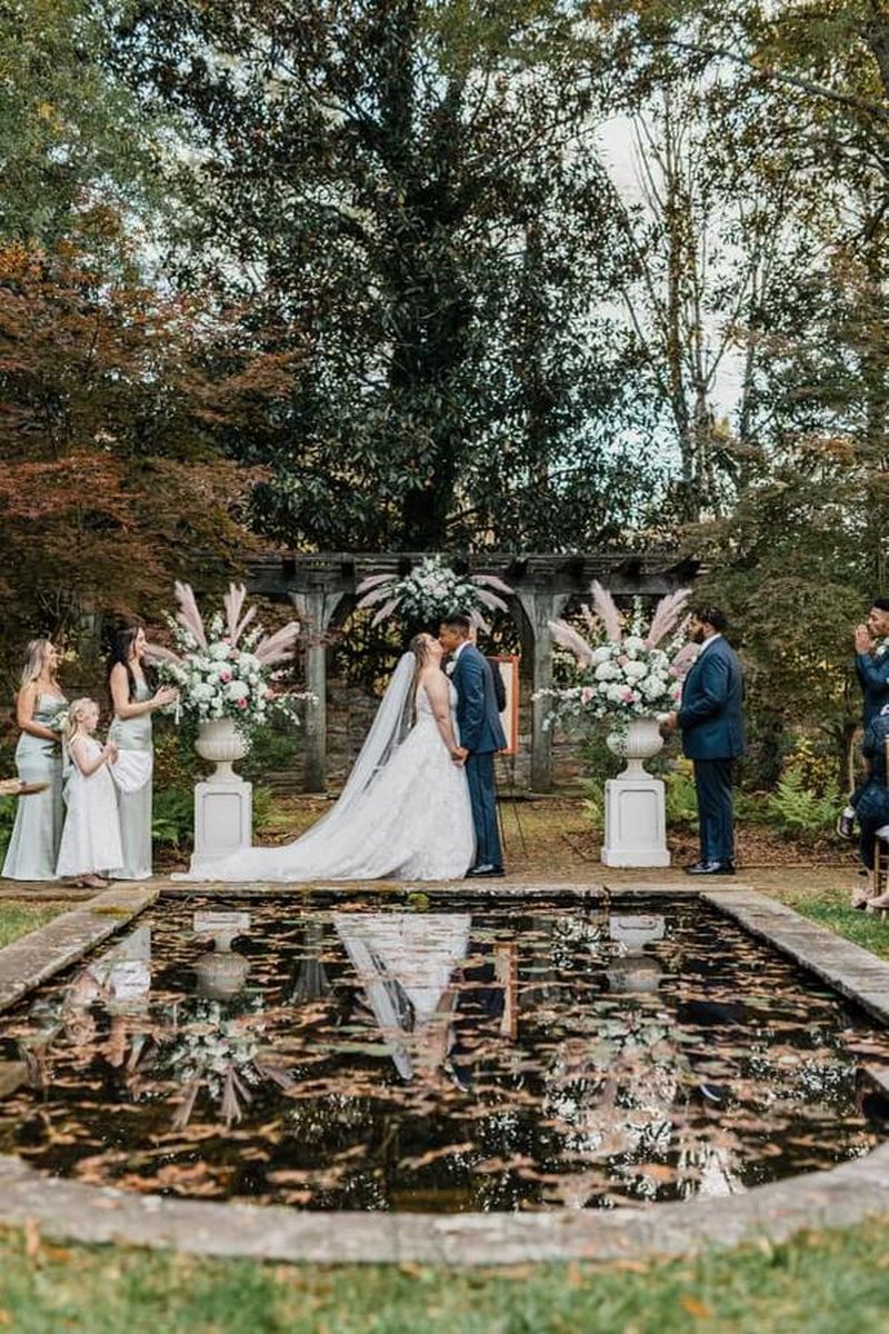 Bride and groom kissing by reflecting pool with stone pergola and pampas grass in autumn