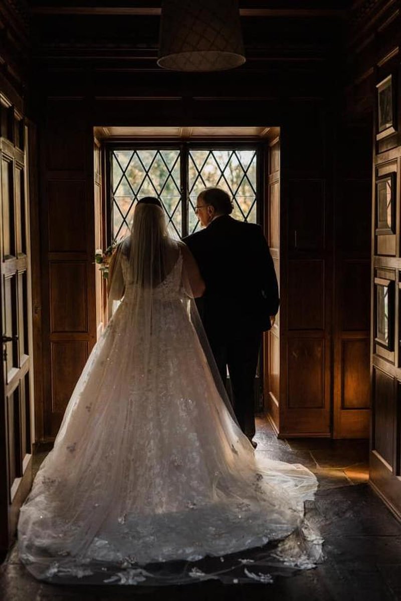 Bride and father silhouetted in dark wood-paneled hallway with diamond-paned window