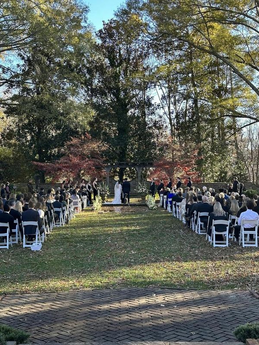 Wide view of outdoor wedding ceremony from back with guests in white chairs toward stone pergola