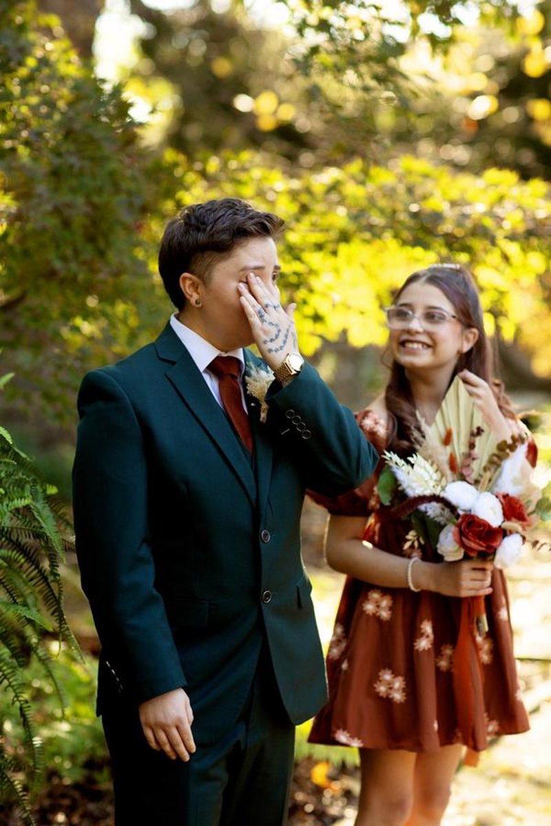 Emotional person in green suit wiping tears while bridesmaid smiles behind them