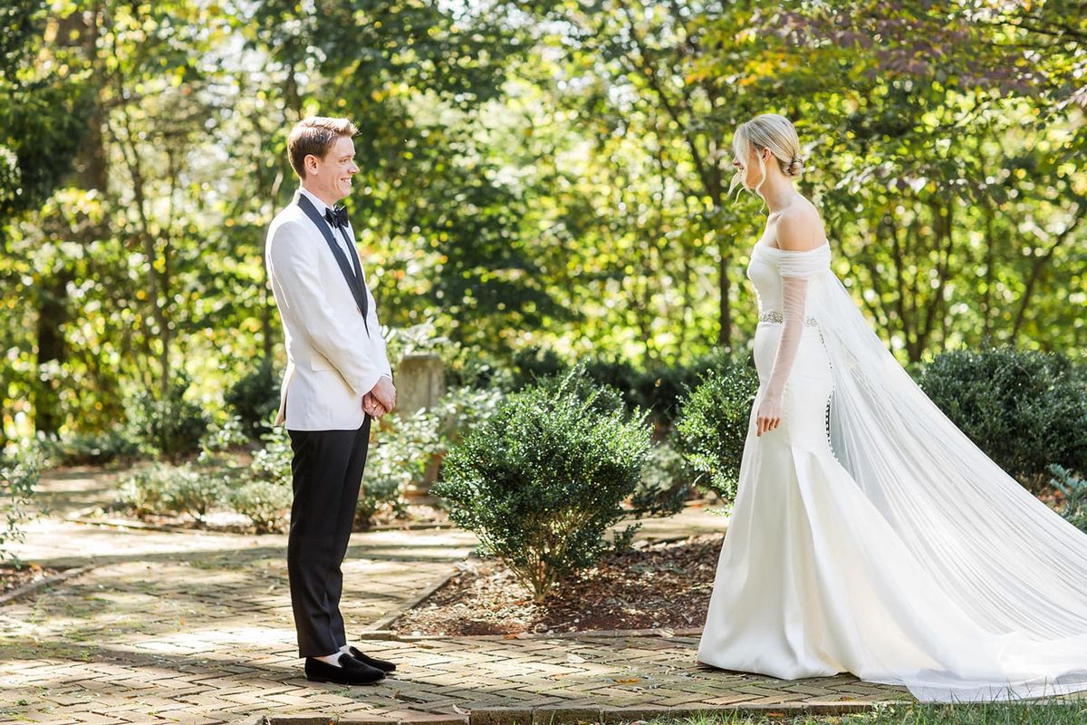 First look moment — groom in white tuxedo sees bride approaching on brick garden pathway